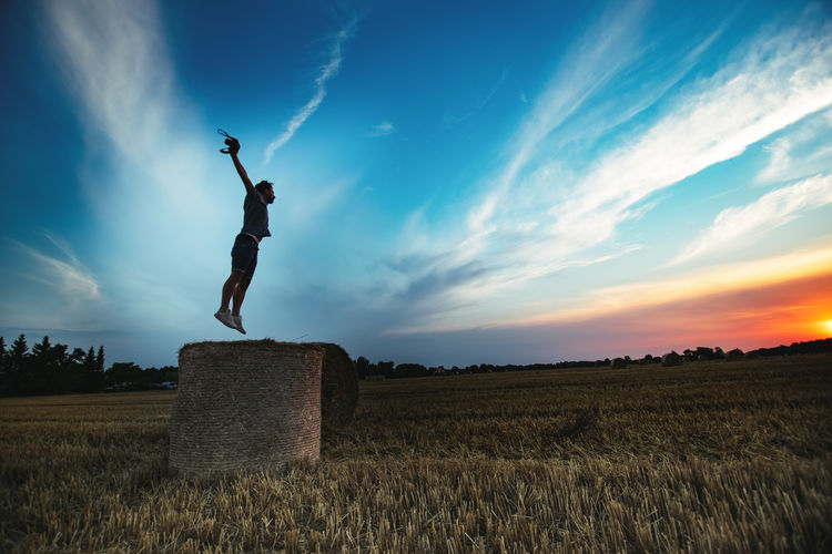 Man jumping on hay bale against dramatic sky during sunset