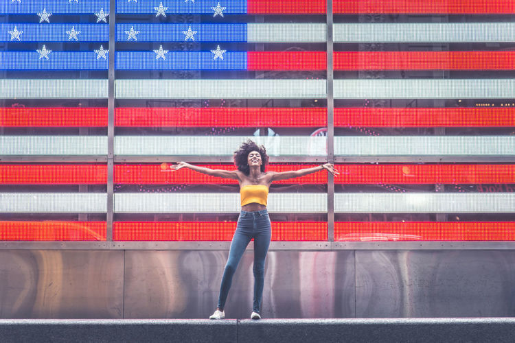 Full length of young woman standing with arms outstretched against neon american flag in city