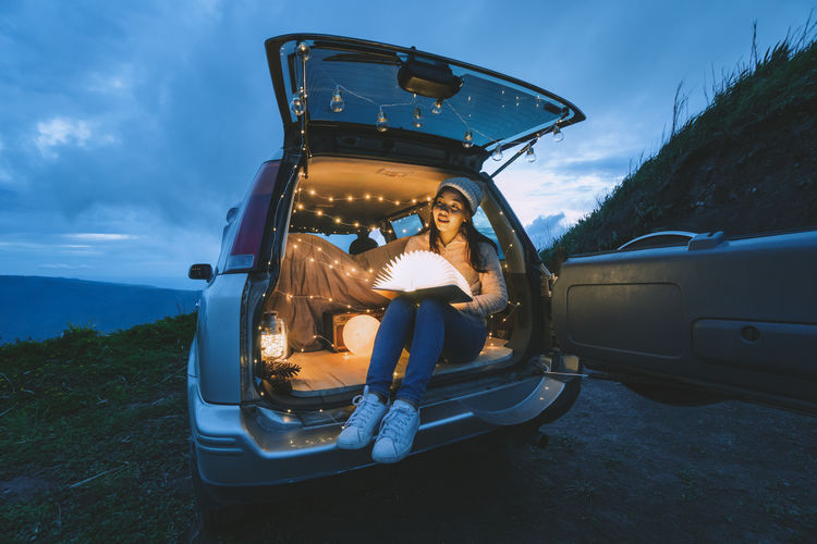 Full length of young woman sitting in illuminated car trunk at dusk