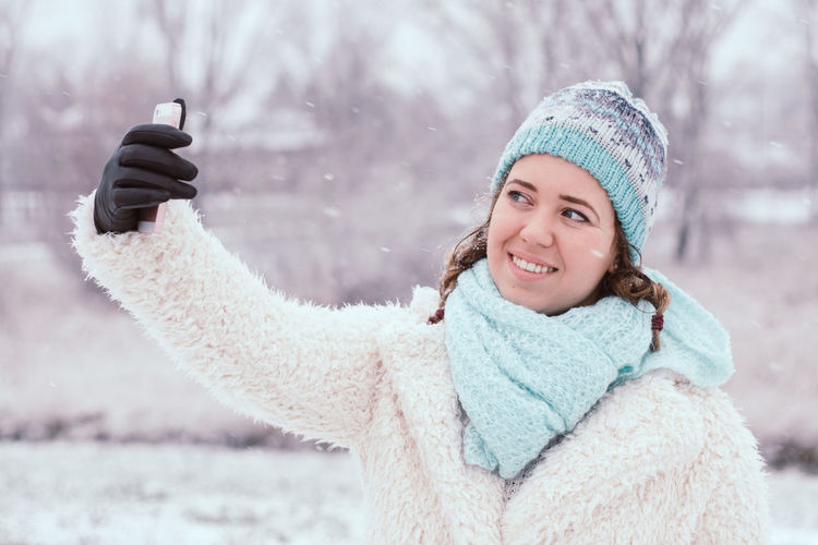 Smiling young woman taking selfie while standing at park during winter