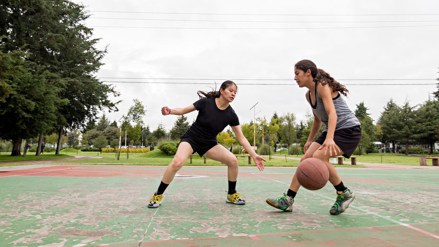 Full body hispanic female athlete blocking opponent dribbling ball during basketball match on cloudy summer day in park