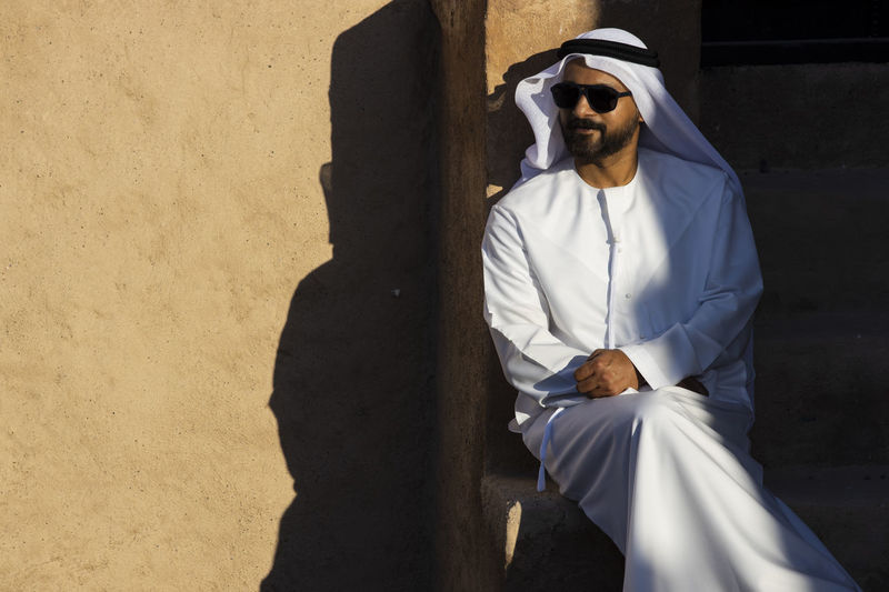Man wearing traditional clothing and sunglasses sitting against wall