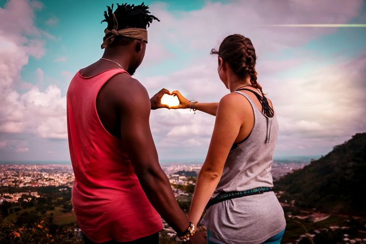 Rear view of man and woman making heart shape with hands while standing on mountain against sky