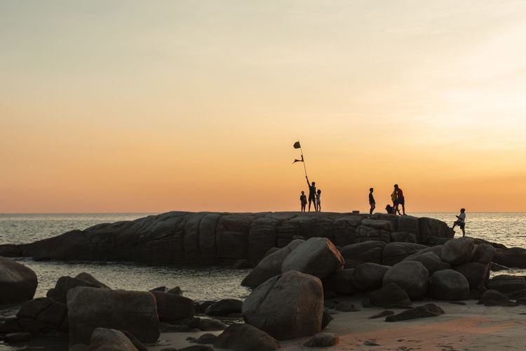 People on rocks by sea against sky during sunset