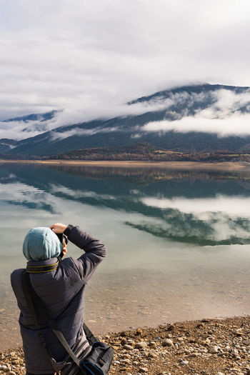 Unrecognizable teenage wearing warm clothes taking picture of the reflected mountain and cloud