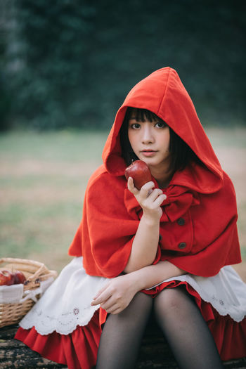 Portrait of beautiful young woman in red riding hood costume eats apple in forest