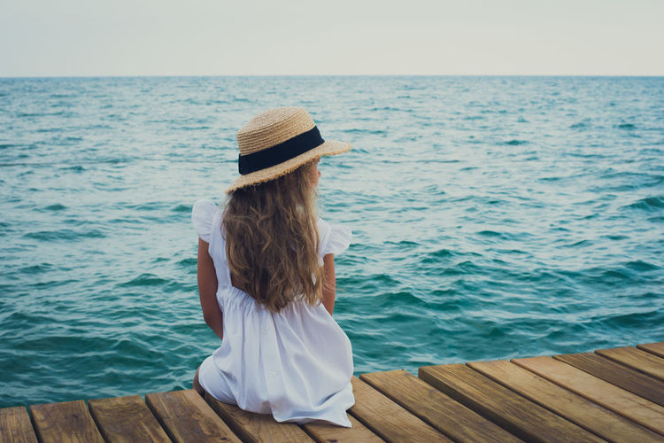 Child in straw hat on sea shore, look from behind