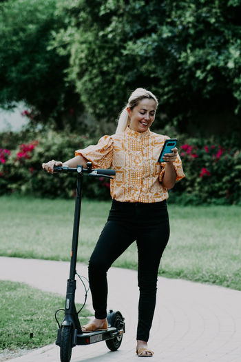 Portrait of a smiling young woman standing against plants with electric scooter 