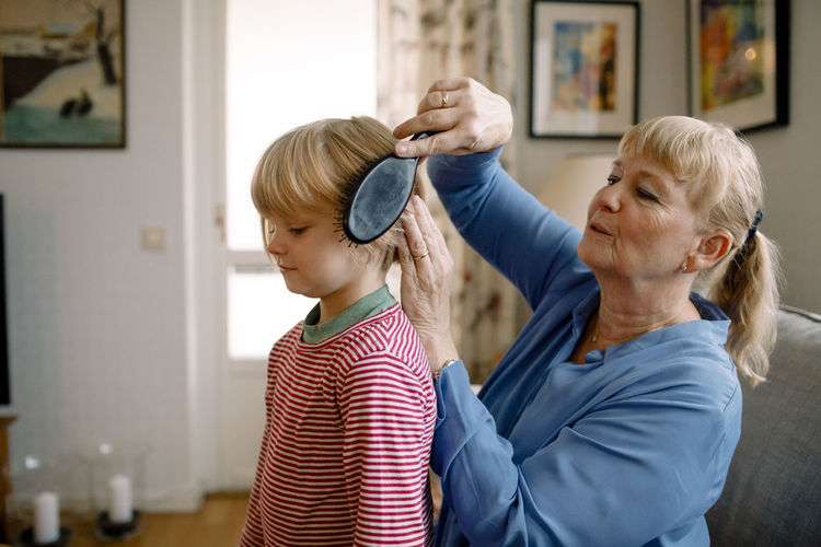 Side view of senior woman combing hair of grandson in living room at home