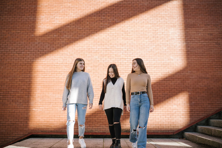 Smiling female friends walking on footpath against brick wall in city