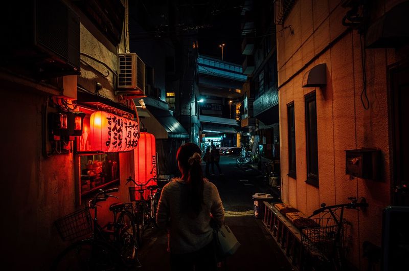 Rear view of woman walking in illuminated alley amidst buildings at night