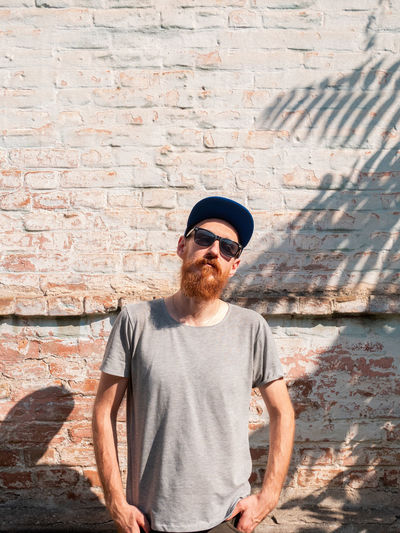 Handsome hipster man with lush beard and mustache on light brick wall background palm leaves shadow.