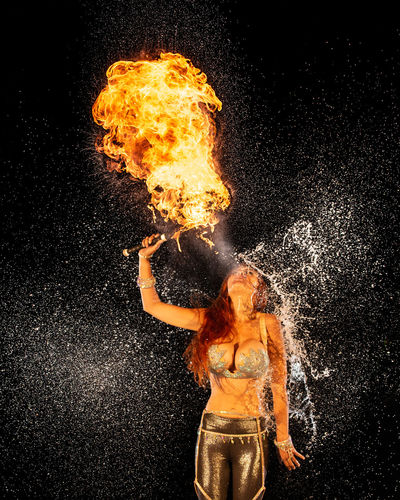 Fire breather donia serena on the beach. while breathing fire, water is thrown at her back.