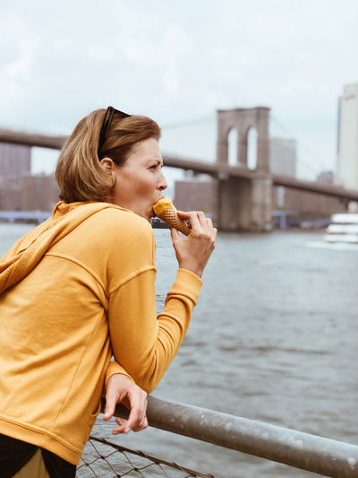 Tourist woman eating  cone of sorbet against background of manhattan city and brooklyn bridge.