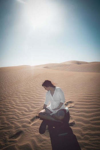 Mid adult woman sitting on sand dune in desert against sky during sunny day