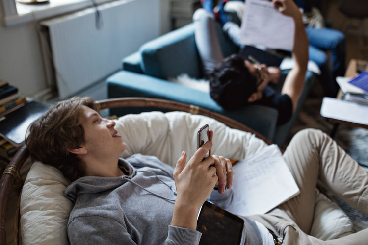 Distracted teenage boy using social media on mobile phone while studying with friend at home
