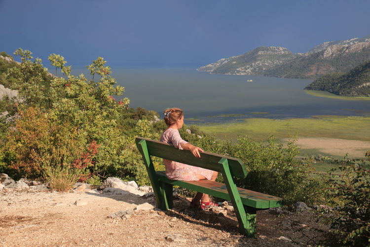 Side view of woman sitting on bench by sea against sky