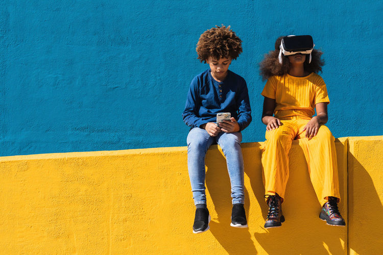 Low angle of african american teen boy with smartphone and girl in vr headset sitting together on colorful wall on street