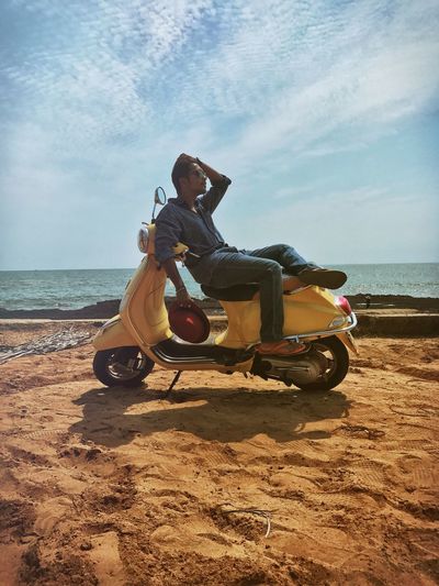 Young man sitting on motor scooter parked at beach against sky
