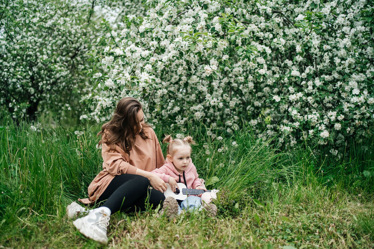 Young mother and baby daughter play the ukulele in a blooming apple orchard in nature