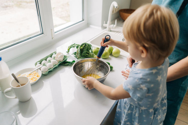 Toddler little daughter helps her mother beat eggs for an omelet.