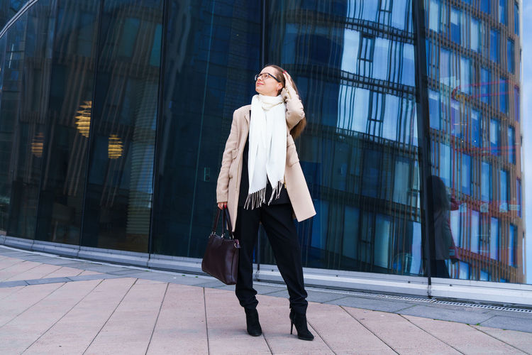 Successful woman with glasses stands in front of an office building with bag
