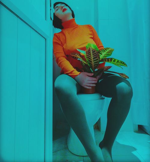 Young woman with potted plant sitting on toilet bowl in bathroom