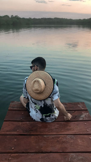 Rear view of man sitting on pier over lake against sky