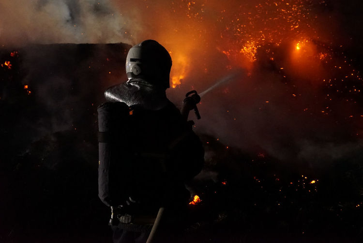 Rear view of firefighter extinguishing fire at night