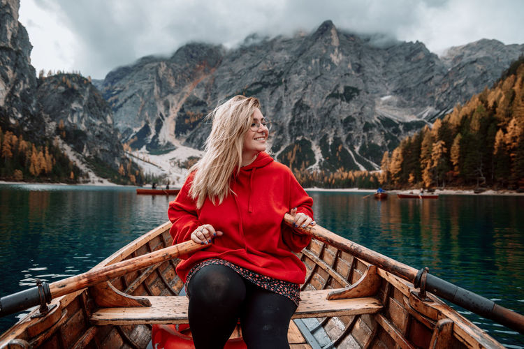 Smiling woman on wooden boat with paddles floating on turquoise water of calm lake on background of majestic landscape of highlands in dolomites in italy alps