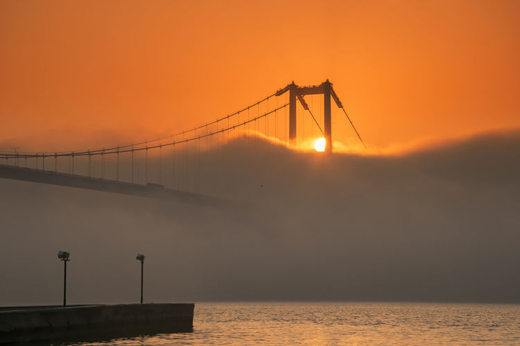 Silhouette bridge over sea against orange sky and fog