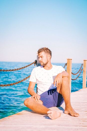 Thoughtful young man looking away while sitting on pier over sea against clear sky
