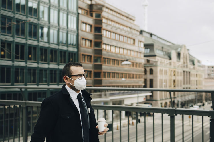 Male entrepreneur with coffee cup by railing in city during pandemic