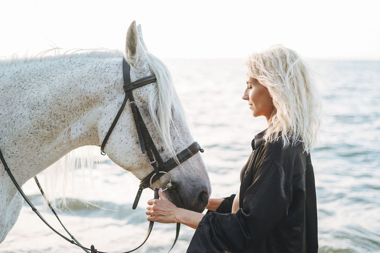 Young blonde woman in black clothes riding white horse on seascape background