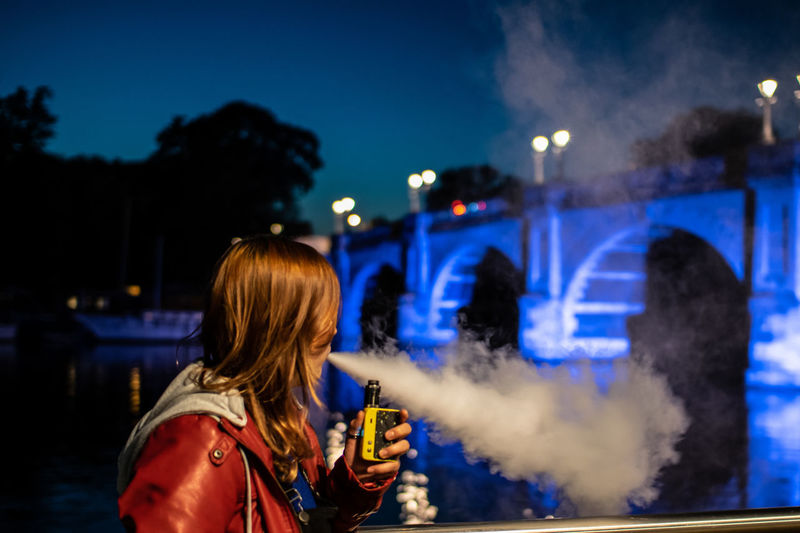 Side view of woman smoking electric cigarette in illuminated lights at night