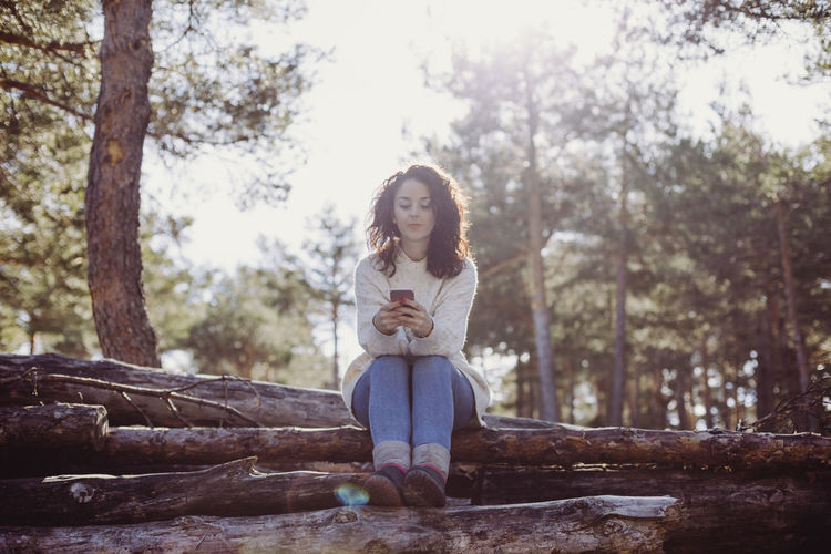 Full length of woman with phone sitting against trees