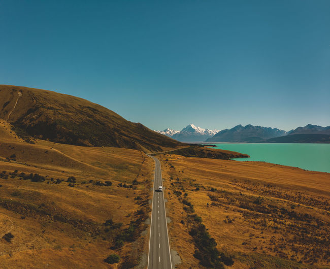 Scenic view of road by mountains and lake against clear blue sky