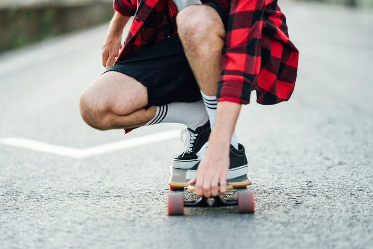 Unrecognizable male hipster in plaid shirt sitting on longboard and riding along road