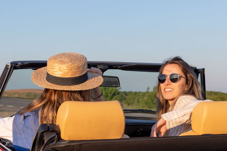 Close up of two young women driving a convertible car while looking at camera on a sunny 