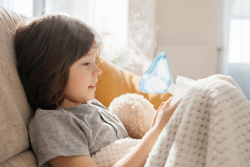 Close-up of a boy doing inhalation at home sitting on the couch with a nebulizer and a mask. 