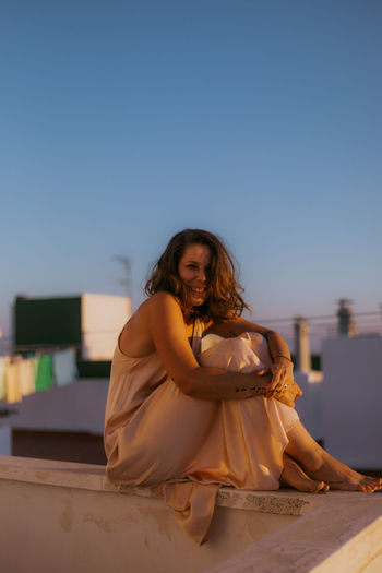 Cheerful happy woman in stylish pink silk dress sitting on fence of balcony with arms around legs and looking at camera during summer sunset