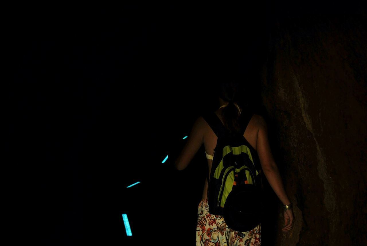 REAR VIEW OF WOMAN STANDING AGAINST ILLUMINATED DARK