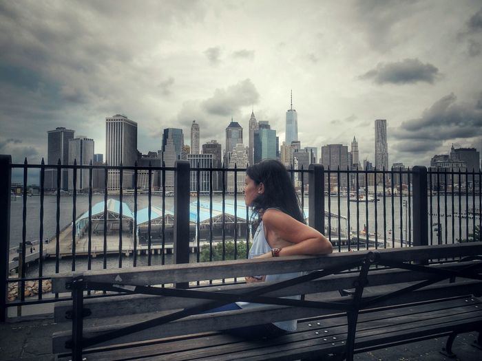 Side view of young woman sitting on bench against skyscrapers in city
