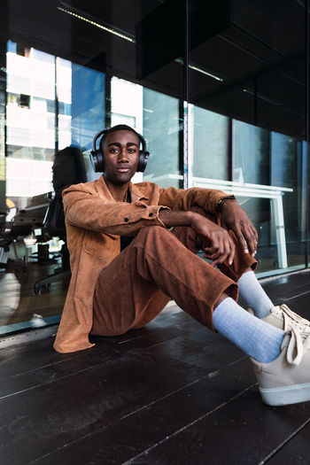 Stylish african american male listening to music in wireless headphones and looking at camera while sitting near glass wall in city