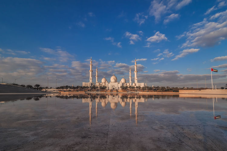 Grand mosque reflecting on calm lake against cloudy sky