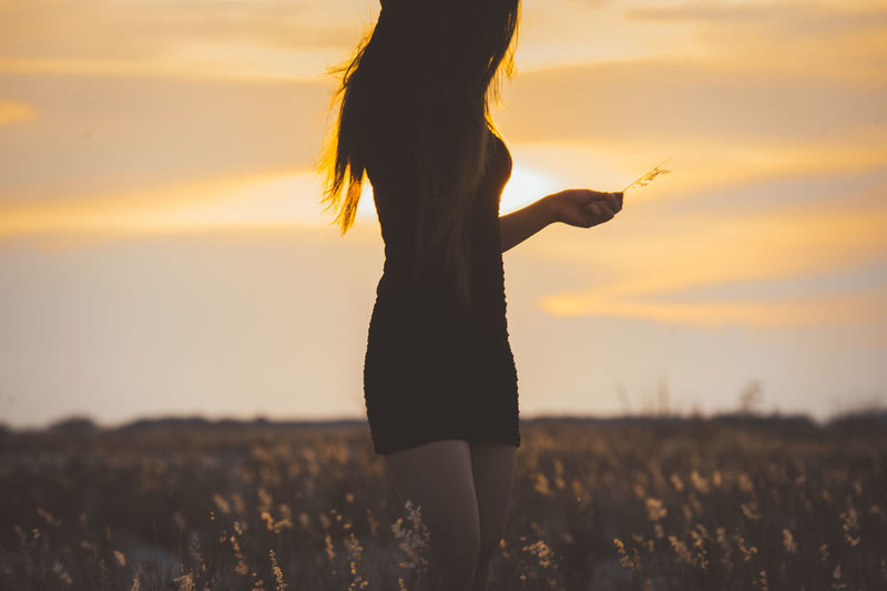 Midsection of woman standing amidst field against sky during sunset
