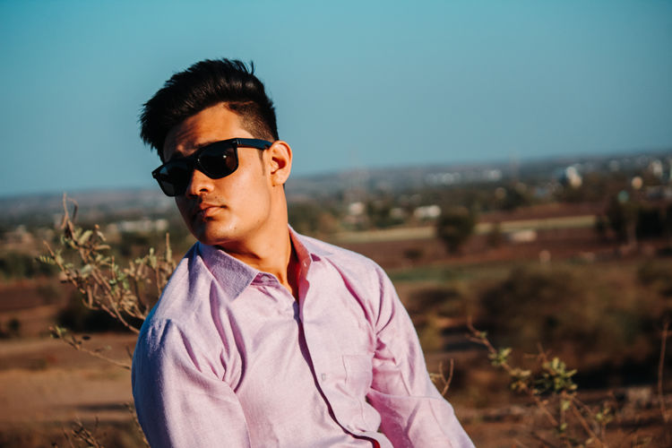 Young man in sunglasses posing while sitting outdoors against sky