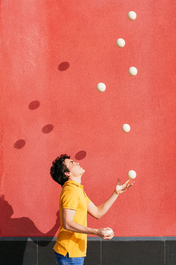 Side view of young talented male performing trick with juggling balls while standing on pavement near bright red wall