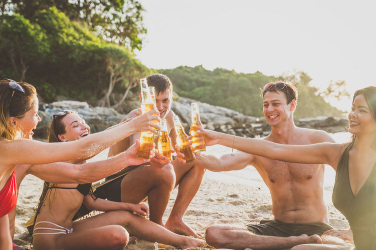Friends toasting beer bottles while sitting at beach