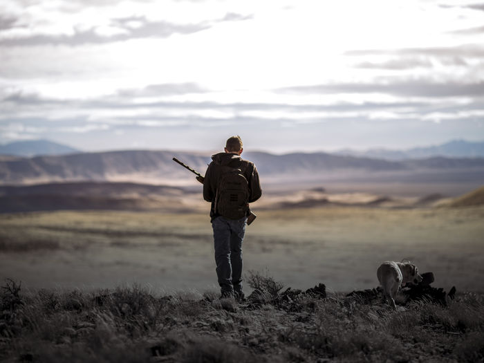 Full length of man with rifle standing on landscape against sky